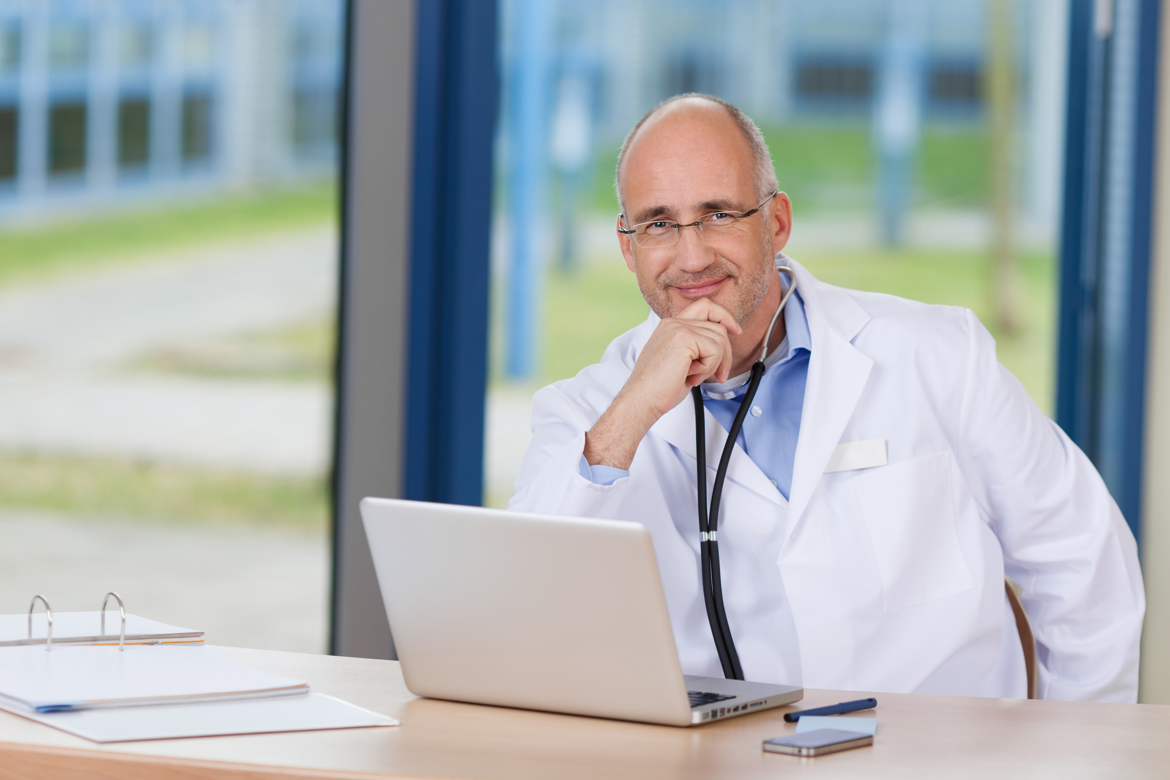 Male Doctor With Hand On Chin And Laptop On Desk